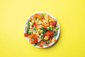 Chopped frozen vegetables in a plate on a yellow background. Corn peas pepper carrots. copy space. photo