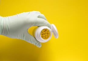 Hand in latex glove holding the plastic container of pills on bright yellow background. photo
