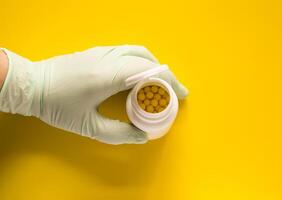 Hand in latex glove holding the plastic container of pills on bright yellow background. photo
