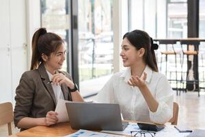 Young businesswoman and colleague collaboration in a modern office environment, discussing ideas and working together on a project with a laptop, documents, and charts, fostering teamwork and photo