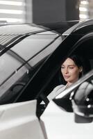Portrait of gorgeous brunette woman make purchase in car showroom. Female sits inside of new, modern, electric car and look at sales agent. Concept for car rental or sale. Test drive. photo