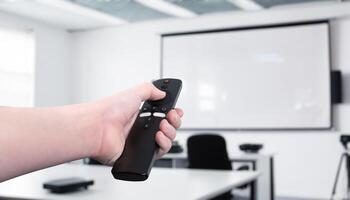 Hand holding a remote control, operating a projector in a conference room or classroom. Concept of presentation control, business meeting, education, technology photo
