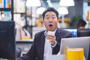 Surprised Man Sitting at Desk in Front of Computer is Holding Cup of Coffee photo