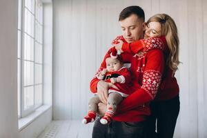 Portrait of happy family mom dad and little daughter in red traditional christmas clothes spending time together in light wooden room near window, copy space photo