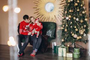 Happy family mom dad and little daughter in red traditional Christmas clothes spending time together near Christmas tree, copy space photo