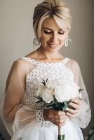 Beautiful blonde bride smiling wearing white wedding dress and holding bouquet indoors. photo