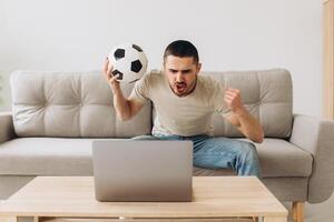 A man sits on a sofa in the living room holding a ball and cheers for the football team watching a match on a laptop photo