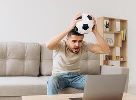 A man sits on a sofa in the living room holding a ball and cheers for the football team watching a match on a laptop photo