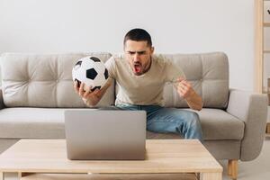 A man sits on a sofa in the living room holding a ball and cheers for the football team watching a match on a laptop photo