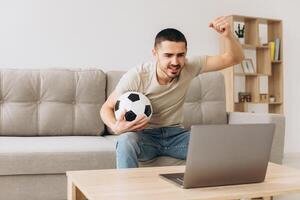 A man sits on a sofa in the living room holding a ball and cheers for the football team watching a match on a laptop photo