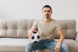 A man sits on a sofa in the living room holding a ball and cheers for the football team and watching a match photo