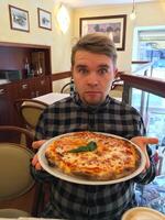 A young funny man with an astonished expression is holding a freshly baked pizza on a white plate in an indoor restaurant setting. Culinary Experience and Authenticity concept photo