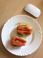 Fresh avocado and smoked salmon toast on white plate. Healthy breakfast standing on a wooden table with a computer mouse nearby. photo