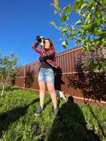 Millennial woman taking photo in a sunny backyard with a small tree and pet dog. Bright and clear blue sky providing a perfect backdrop for outdoor photography. Photographer, connection with nature
