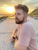 Bearded man gazes thoughtfully at the sunset, leaning against a concrete railing on a scenic promenade. Soft glow of the setting sun creates a serene and reflective atmosphere photo