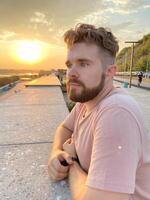 Bearded man gazes thoughtfully at the sunset, leaning against a concrete railing on a scenic promenade. Soft glow of the setting sun creates a serene and reflective atmosphere photo