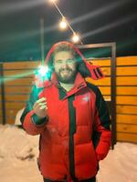 Bearded man in a red jacket holds a glowing sparkler, standing outside on a snowy winter night, with festive string lights hanging overhead. Christmas and New Year celebration concept photo