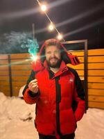 Bearded man in a red jacket holds a glowing sparkler, standing outside on a snowy winter night, with festive string lights hanging overhead. Christmas and New Year celebration concept photo