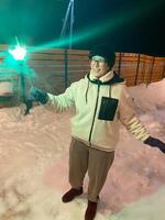 Woman in a red jacket holds a glowing sparkler, standing outside on a snowy winter night, with festive string lights hanging overhead. Christmas and New Year celebration concept photo