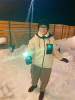 Woman in a red jacket holds a glowing sparkler, standing outside on a snowy winter night, with festive string lights hanging overhead. Christmas and New Year celebration concept photo