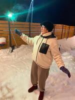 Woman in a red jacket holds a glowing sparkler, standing outside on a snowy winter night, with festive string lights hanging overhead. Christmas and New Year celebration concept photo