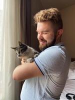Millennial man gently holds a cat by a sunlit window, capturing a moment of serene companionship. Warm light, sense of peace and connection between human and pet. photo
