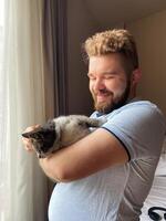 Millennial man gently holds a cat by a sunlit window, capturing a moment of serene companionship. Warm light, sense of peace and connection between human and pet. photo