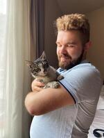 Millennial man gently holds a cat by a sunlit window, capturing a moment of serene companionship. Warm light, sense of peace and connection between human and pet. photo
