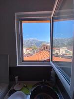 View of distant mountains framed perfectly by an open window, revealing rooftops and a clear blue sky. Indoor sink area contrasts with the vibrant outdoor scenery. photo