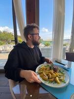 Bearded man enjoys a meal at a seaside restaurant, with a plate of steak and potatoes. Bright and open atmosphere, surrounded by palm trees and a scenic view of the beach outside photo