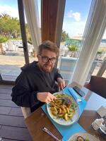 Bearded man enjoys a meal at a seaside restaurant, with a plate of steak and potatoes. Bright and open atmosphere, surrounded by palm trees and a scenic view of the beach outside photo