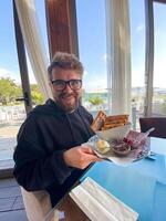 Man smiles while holding a plate of waffles with sides of ice cream, berries, and chocolate at a seaside restaurant. Bright and sunny setting, with palm trees visible outside, photo