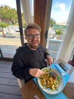 Bearded man enjoys a meal at a seaside restaurant, with a plate of steak and potatoes. Bright and open atmosphere, surrounded by palm trees and a scenic view of the beach outside photo