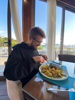 Bearded man enjoys a meal at a seaside restaurant, with a plate of steak and potatoes. Bright and open atmosphere, surrounded by palm trees and a scenic view of the beach outside photo