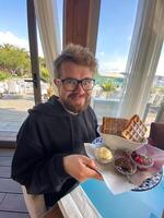 Man smiles while holding a plate of waffles with sides of ice cream, berries, and chocolate at a seaside restaurant. Bright and sunny setting, with palm trees visible outside, photo