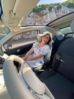 Cheerful woman is sitting in a car with the roof open, enjoying a sunny day. Wearing a summer hat and laughing with joy photo