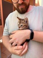 Millennial man in a straw hat and sunglasses holds lovingly at a small kitten in his hands. Tenderness moment captures the bond between human and pet. Themes of care and affection photo