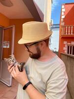 Millennial man in a straw hat and sunglasses holds lovingly at a small kitten in his hands. Tenderness moment captures the bond between human and pet. Themes of care and affection photo