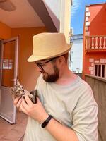 Millennial man in a straw hat and sunglasses holds lovingly at a small kitten in his hands. Tenderness moment captures the bond between human and pet. Themes of care and affection photo
