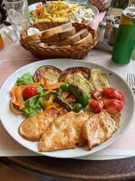 Delicious meal featuring grilled vegetables and chicken breast is elegantly presented on a white plate. Basket of sliced bread and a soft drink can, creating a cozy dining atmosphere photo