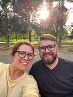 Joyful couple enjoys a sunny day outdoors, smiling brightly as they take a selfie in park. Warm sunlight filters through the trees, highlighting the happiness and connection shared between them photo