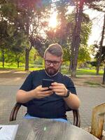 Bearded man sits in a park, focused intently on smartphone with sun shining through the trees in background. Peaceful setting highlights the blend of technology photo