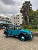 Classic vintage car rests in an urban setting, juxtaposing modern architecture with nostalgic charm. Bright teal vehicle stands out against backdrop of palm trees photo