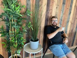 Bearded man in casual attire sits on a chair, holding a cup of coffee, against a stylish wooden wall with potted plants. Relaxed and modern atmosphere, perfect for enjoying a peaceful moment photo
