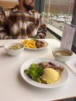 Millennial man in a plaid shirt sits at a white table with a hearty meal in front of him, featuring mashed potatoes, broccoli and big portion of protein. photo
