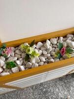 Wooden planter filled with white pebbles and colorful artificial flowers decorates an interior space. Arrangement adds a touch of nature with minimal maintenance required photo