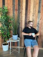Bearded man in casual attire sits on a chair, holding a cup of coffee, against a stylish wooden wall with potted plants. Relaxed and modern atmosphere, perfect for enjoying a peaceful moment photo