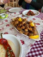 Table set for a cozy Balkan meal with golden fried fish garnished with lemon wedges. Red peppers and a glass of wine complete inviting dining scene in a charming rustic restaurant photo