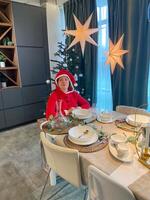Millennial woman in a Santa suit sits thoughtfully and sad at a festive dining table, surrounded by Christmas decorations and a lit tree. holiday reflection and problem loneliness in holidays photo