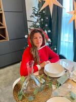 Millennial woman dressed in a Santa outfit sits at a beautifully set holiday table, surrounded by festive decorations and a Christmas tree. Cozy scene illuminated photo